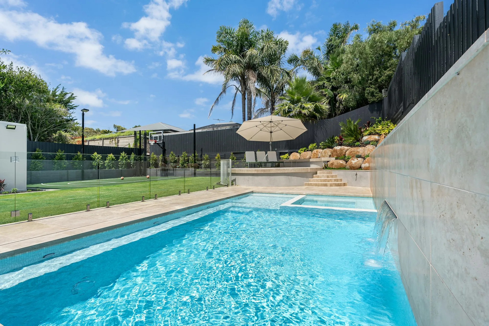 Swimming pool with a glass panel barrier on the lawn side and a dark board fence behind it
