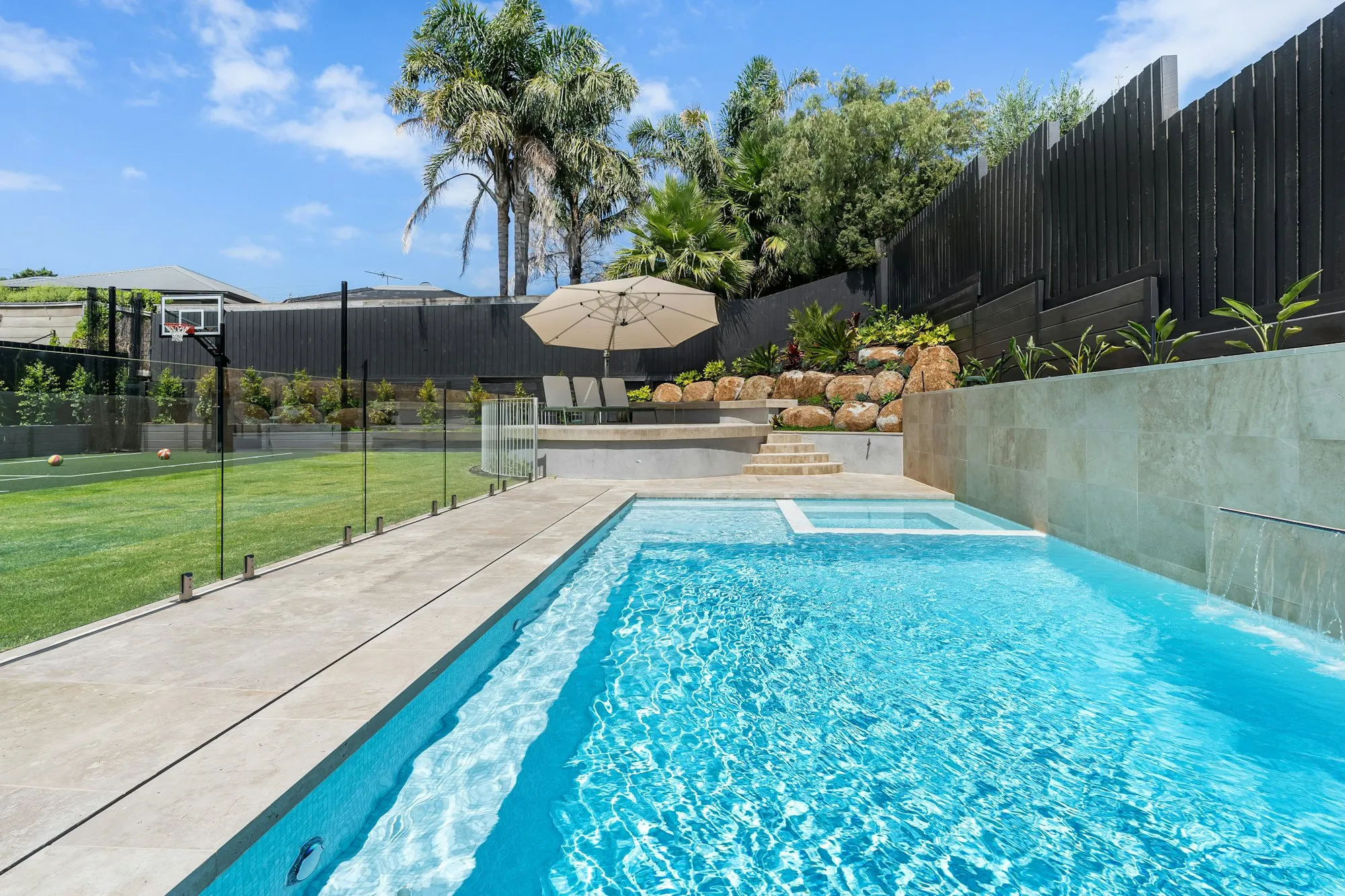Swimming pool enclosed by a glass panel barrier, with a dark wooden fence along the back of the yard