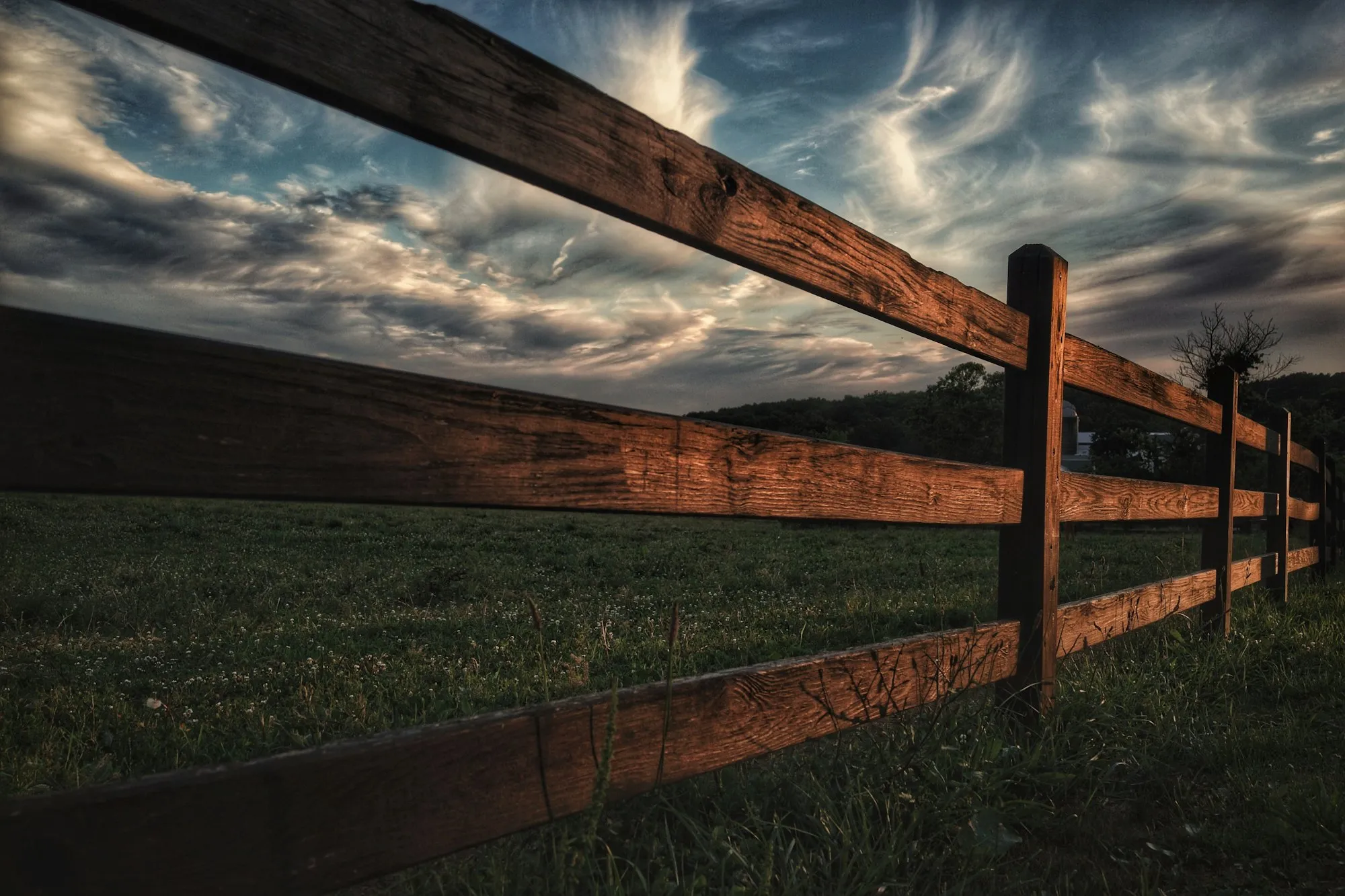 Dark wooden three-rail fence crossing a field under a streaked evening sky